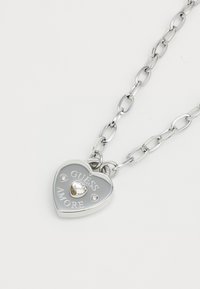 Silver chain necklace with a heart-shaped pendant featuring "GUESS AMORE" engraving, a clear crystal heart, and two accent stones.