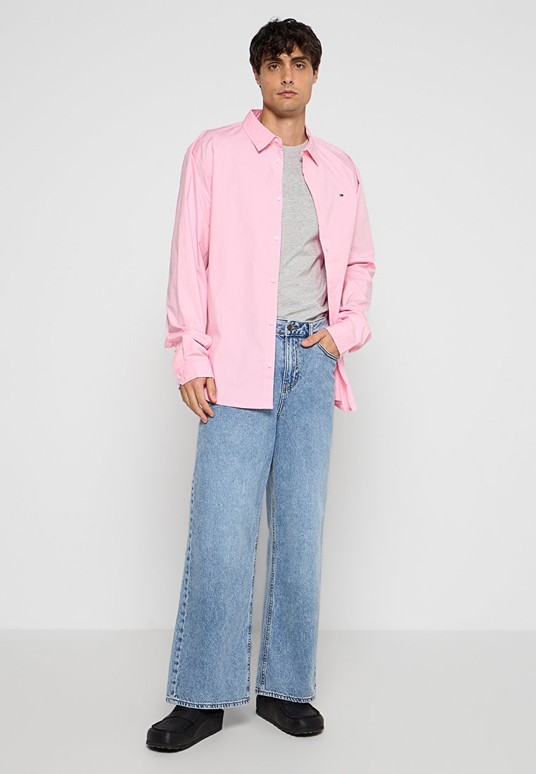 Rosa Langarm-Hemd mit Knöpfen über einem grauen T-Shirt, kombiniert mit weit geschnittenen blauen Jeans und schwarzen Schuhen. Schlichtes Design, lockere Passform.