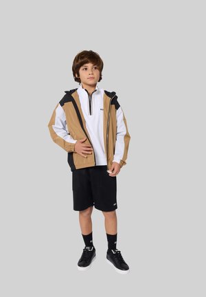 Veste à capuche marron et blanche sur un haut zippé blanc, associée à un short noir et des baskets noires assorties. Tissu lisse avec des détails zippés.