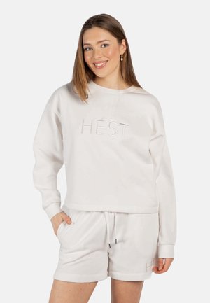 Kvinne som smiler, iført hvit hettegenser og matchende shorts med "HÉST" brodert, stående med den ene hånden i lommen mot lys bakgrunn.