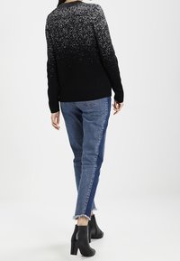 Frau trägt einen schwarz-weiß gesprenkelten Pullover, zweifarbige blaue Jeans mit ausgefransten Säumen und schwarze Stiefeletten mit Absatz, steht mit dem Rücken zur Kamera.