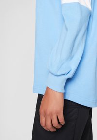 Ljusblå långärmad skjorta med ribbade muddar; har en vit snedställd detalj. Tyget verkar mjukt med en något strukturerad yta.