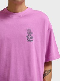 Mężczyzna noszący fioletową koszulkę z małą grafiką deskorolkarza i napisem „hummel SPORTSWEAR” na piersi, widoczny tatuaż na przedramieniu.