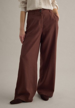 Vrouw draagt een beige shirt met lange mouwen en een hoge taille, wijd uitlopende bruine broek, staat met één hand in haar zak.