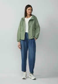 Veste zippée vert clair avec capuche, dotée de deux poches avant et de poignets élastiques, portée sur un haut blanc et un jean bleu à jambes larges.