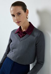 Grijze v-hals trui met lange mouwen, gedragen over een donkerburgundy overhemd met kraag. De textuur van de stof heeft een geribbelde hals.