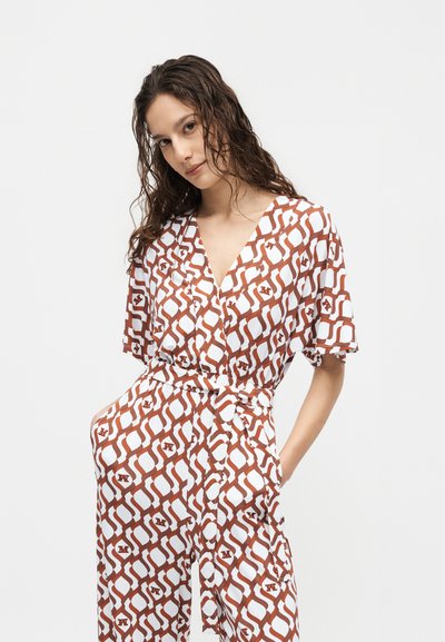 Γυναίκα με μακριά, κυματιστά μαλλιά φοράει ολόσωμη φόρμα με γεωμετρικό σχέδιο σε λευκό και καφέ χρώμα, με ζώνη, κοντά μανίκια και τα χέρια στις τσέπες.