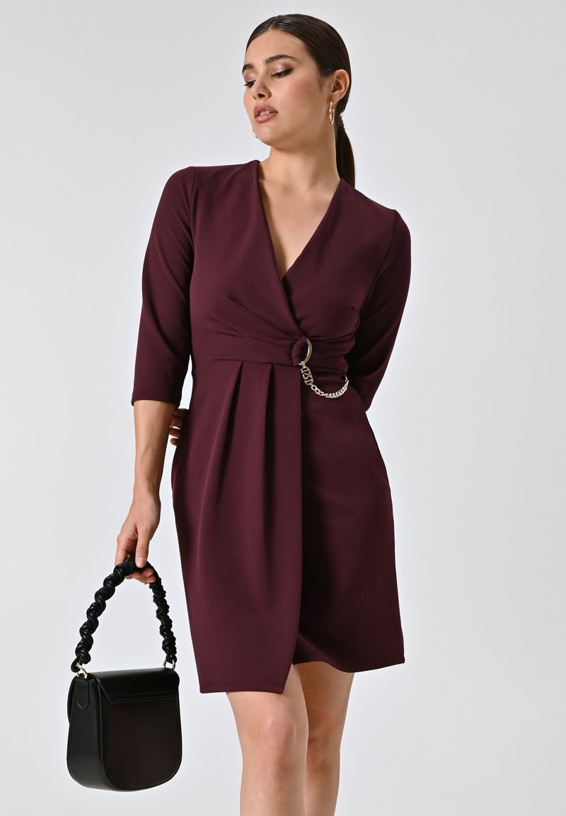Burgunderfarbenes V-Ausschnittkleid mit 3/4-Ärmeln, verdrehtem Vorderteil und Kettenakzent. Das Modell hält eine schwarze Tasche mit geflochtenem Griff.
