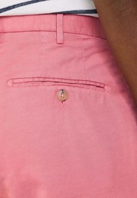Helle rosa Baumwollshorts mit einer Gesäßtasche, die einen runden Knopfverschluss und eine flache Paspelierung aufweist. Der Stoff hat eine glatte Textur.