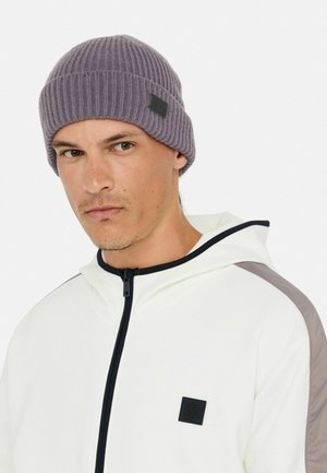 Berretto lavorato a maglia con texture a costine morbide, colore lilla chiaro, con una piccola etichetta nera. Indossato con una felpa bianca con zip.