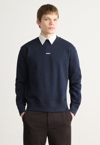Marineblå genser med V-hals design og ribbede mansjetter. Har "BOSS" logo i hvitt. Brukt over en hvit skjorte med krage.