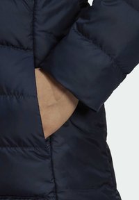 Main à l'intérieur de la poche latérale d'une veste matelassée bleu foncé montrant un gros plan du tissu et des détails de couture.