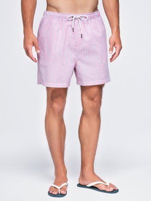 Mann trägt rosa-weiß gestreifte Badeshorts mit Tunnelzug in der Taille und weiße Flip-Flops vor weißem Hintergrund.