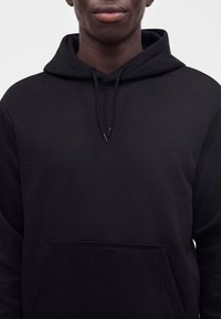 Person trägt einen schlichten schwarzen Kapuzenpullover mit Kordelzug und Bauchtasche, gezeigt von der Brust bis zum Kinn vor hellem, einfarbigem Hintergrund.