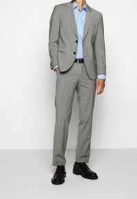 Lichtgrijs, op maat gemaakt pak met een enkelsluitende blazer, gecombineerd met een blauwe blouse en zwarte dresschoenen. Vloeiend stof, scherpe lijnen.