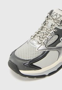 Sneaker grigio con tomaia in mesh e sintetico, accenti lucidi, suola testurizzata e lacci con motivo. Presenta una punta arrotondata e collare imbottito.