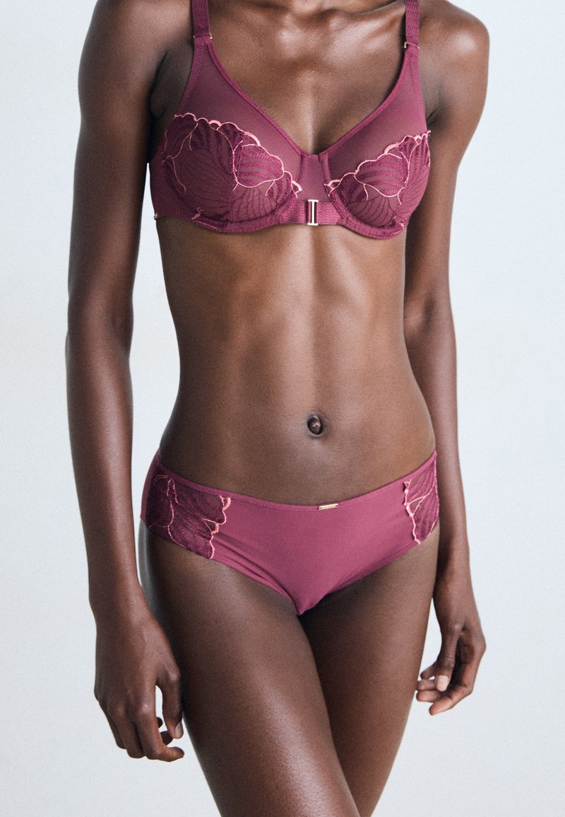 Woman wearing burgundy lace-embellished bra and matching panties, standing against a plain light background.