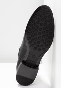 Semelle de botte noire avec motifs de crampons circulaires, taille 37 indiquée, fabriquée en Espagne, reposant contre une surface blanche.