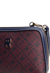 Piros és fekete mintázatú Tommy Hilfiger táska kék bőr díszítéssel, arany cipzárral és arany "TH" logóval az elején.