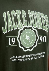 Zielona koszulka bawełniana z dużym białym i jasnoszarym tekstem "JACK & JONES", z okrągłym logo i informacjami o roku założenia.