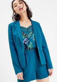 Blazer teal à un bouton et col à revers, superposé à un haut à imprimé floral aux verts et roses vibrants. Un short ajusté complète la tenue.