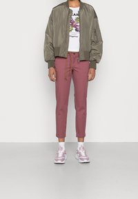 Osoba ubrana w oliwkową bombkę, białą koszulkę z kwiatowym nadrukiem, różowe spodnie z linowym paskiem, białe skarpetki i różowo-białe sportowe buty.