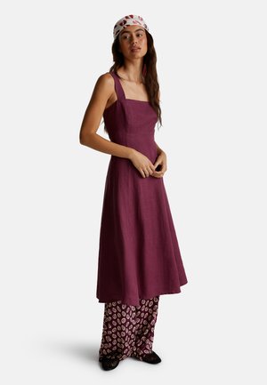 Ein ärmelloses, burgunderfarbenes Leinenkleid mit quadratischem Ausschnitt, kombiniert mit gemusterten, locker sitzenden Hosen in Lila und Creme.