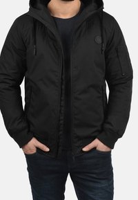 Black hooded jacket in a matte finish, featuring zippered pockets and adjustable drawstrings. Worn over a dark shirt and jeans.