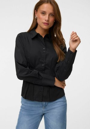 Femme aux longs cheveux bruns portant une chemise noire cintrée à boutons et un jean bleu clair, debout avec un bras croisé et l'autre levé.