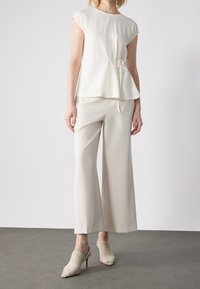 Biały, teksturowany top z ozdobą w postaci kokardy w talii, zestawiony z jasnobeżowymi szerokimi spodniami i kremowymi plecionymi mulesami na obcasie.