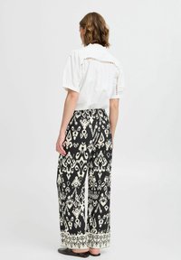 Chemise blanche à manches courtes avec des découpes dans le dos ; pantalon large à motifs noir et crème avec un ourlet décoratif. Chaussures plates.