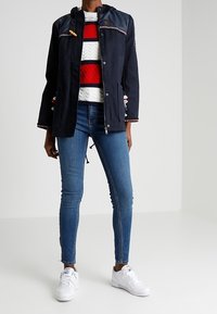 Veste imperméable bleu marine avec capuche, ornée d'un galon décoratif et de cordons de serrage, portée par-dessus un pull à motifs rouge, blanc et bleu, un jean bleu et des baskets blanches.