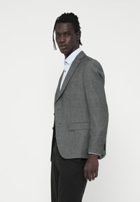 Sivý károvaný blazer s výrezovými klopami, jedným gombíkom a dvomi prednými vreckami. Obláčený cez svetlomodrú košeľu, má textúrovanú tkaninu.