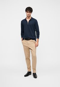 Μπλε σκούρο πόλο μπλουζάκι με μακριά μανίκια και φερμουάρ στον γιακά, συνδυασμένο με μπεζ παντελόνια και μαύρα μποτάκια μέχρι τον αστράγαλο. Λείο ύφασμα, εφαρμοστό σχέδιο, χωρίς σχέδια.