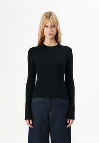 Mulher com cabelo loiro vestindo um suéter justo de manga longa preto canelado e jeans largos de tom azul escuro contra um fundo branco.