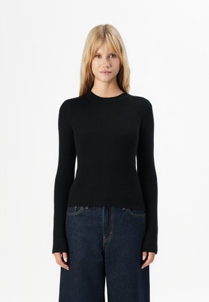 Vrouw met blond haar, gekleed in een nauwsluitende zwarte geribbelde longsleeve trui en donkerblauwe wijde jeans, tegen een witte achtergrond.
