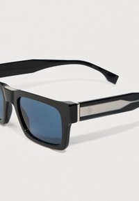 Μαύρα ορθογώνια γυαλιά ηλίου με μπλε φιμέ φακούς και ασημί λεπτομέρειες στα μπράτσα, τοποθετημένα σε μια απλή ανοιχτόχρωμη επιφάνεια.