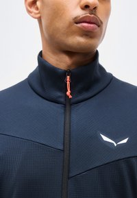 Κοντινό πλάνο ενός ατόμου που φοράει μπλε σκούρο μπουφάν με φερμουάρ και ψηλό γιακά, με λευκό λογότυπο στο στήθος.