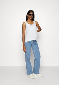 Top branco sem mangas combinado com jeans azul claro. O modelo usa óculos de sol pretos e tênis brancos, posando contra um fundo neutro.