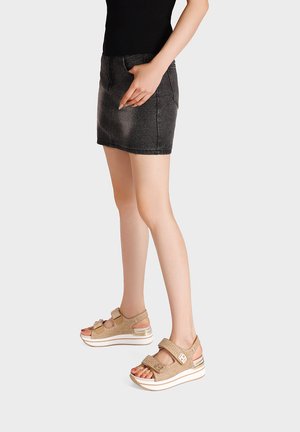 Donna con top nero e minigonna in denim scuro con sandali beige con plateau, in piedi davanti a uno sfondo semplice.