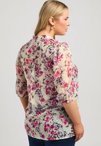 Häikäisevä kukkablouse, jossa on vaalealla taustalla vaaleanpunaisia ja violetteja kukkia, kolmanneksi vartalonpituiset hihat ja rypytetty takaosa.