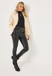 Pantalones de cuero sintético negros con cintura alta y diseño ajustado, combinados con una camisa de pana crema sobre un cuello de tortuga negro acanalado y botas negras con cordones.