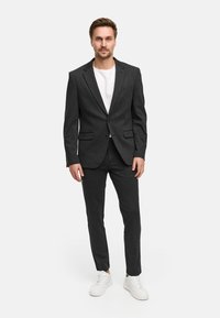 Mørkegrå dress med en ensfarget jakke, kombinert med matchende bukser og hvite joggesko. Begge plaggene er skreddersydd for en tettsittende look.