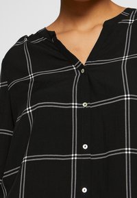 Schwarzes Button-Up-Hemd aus leichtem Stoff mit einem weißen Karomuster, V-Ausschnitt und abgerundetem Saum mit silberfarbenen Knöpfen.