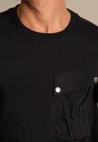 Sudadera negra con cuello redondo que presenta un bolsillo de tela texturizada con botones de presión, aportando contraste y un detalle utilitario al diseño.