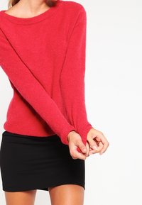 Puszysty czerwony sweter z szerokim dekoltem, długimi rękawami i ściągaczami, w połączeniu z dopasowaną czarną spódnicą. Widoczna jest miękka faktura.