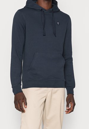 Mand iført en mørkeblå hættetrøje med frontlomme og beige bukser, stående mod en ensartet lys baggrund.