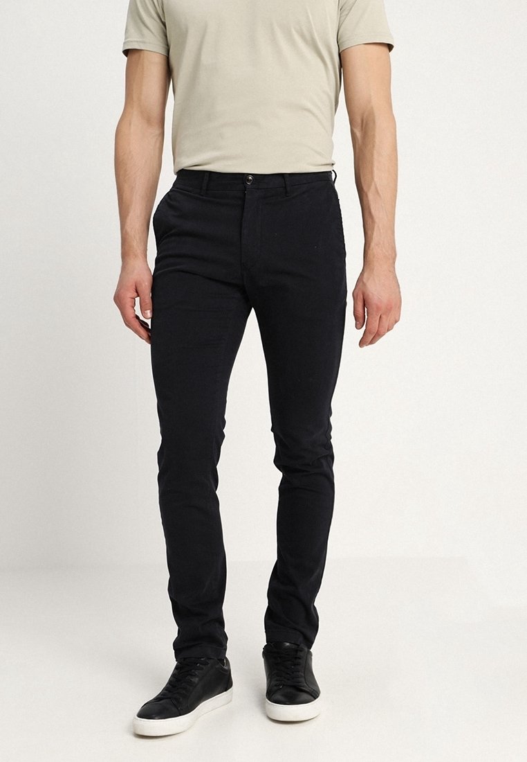 Schwarze Slim-Fit-Hosen aus strukturiertem Stoff mit Knopfverschluss und Gürtelschlaufen, kombiniert mit schwarzen Sneakers und einem hellbeigen T-Shirt.