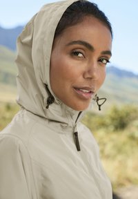 Šviesiai smėlio spalvos liemenė su reguliuojamais raiščiais, lygiu paviršiumi ir minimalistišku dizainu, dėvima modelio lauke su žaliu kraštovaizdžiu fone.