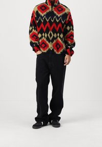 Pullover in fleece con colletto alto, caratterizzato da un motivo geometrico in rosso, nero e beige. Abbinato a jeans neri e sneakers.